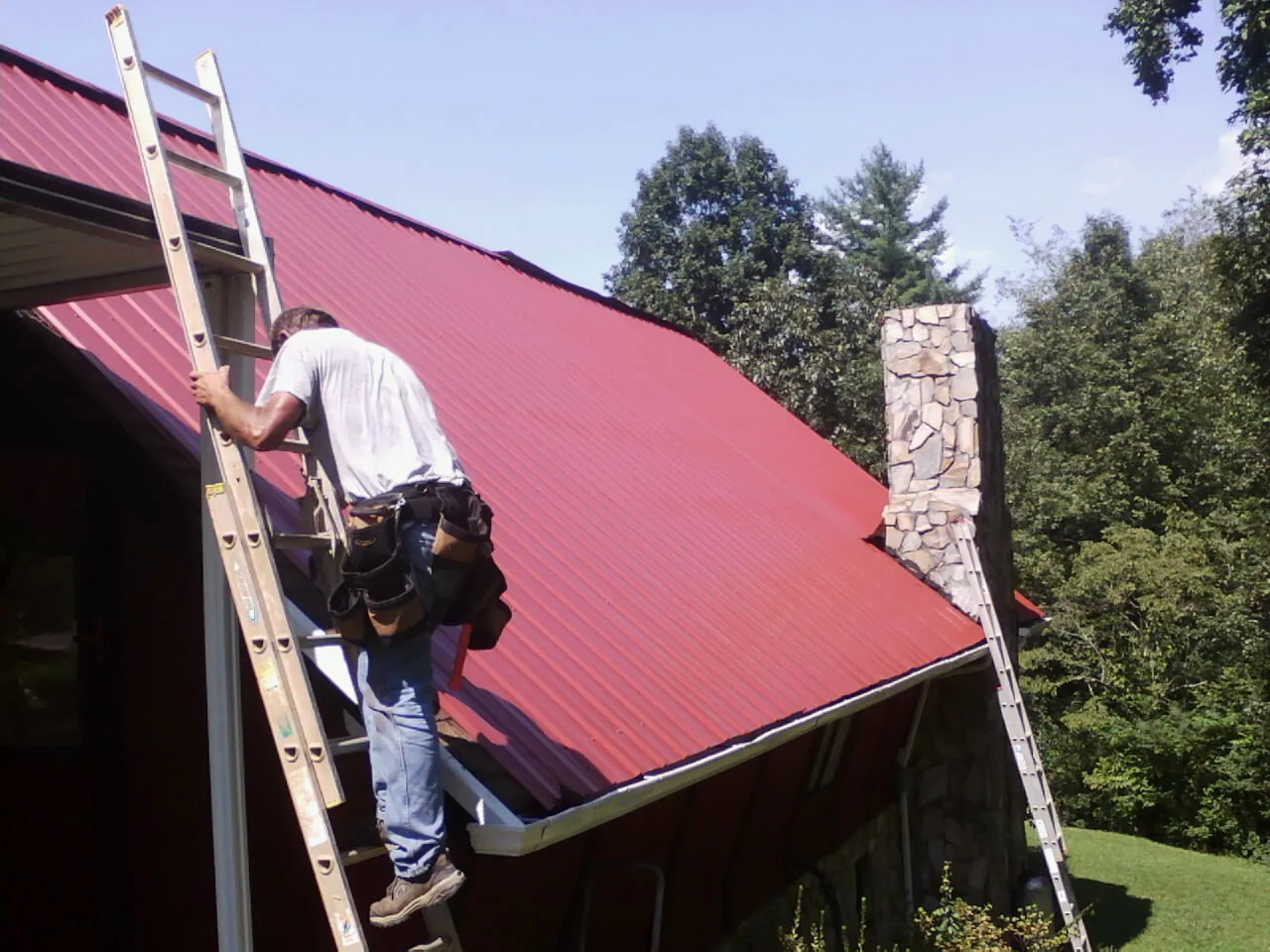 Roofer on a ladder beside a red metal roof and stone chimney — Soffit & Fascia Repair by Roofing Brothers in Panama